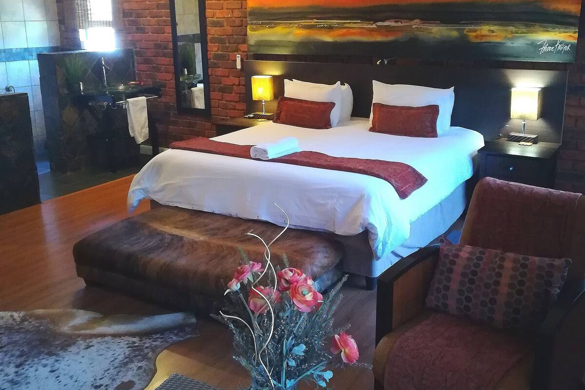 Ein Hotelzimmer mit Backsteinwand, einem großen Bett mit weißer Bettwäsche, einem Kopfteil mit einem Gemälde, einem Nachttisch mit einer Lampe und einem kleinen Tisch mit einer Blumenvase.