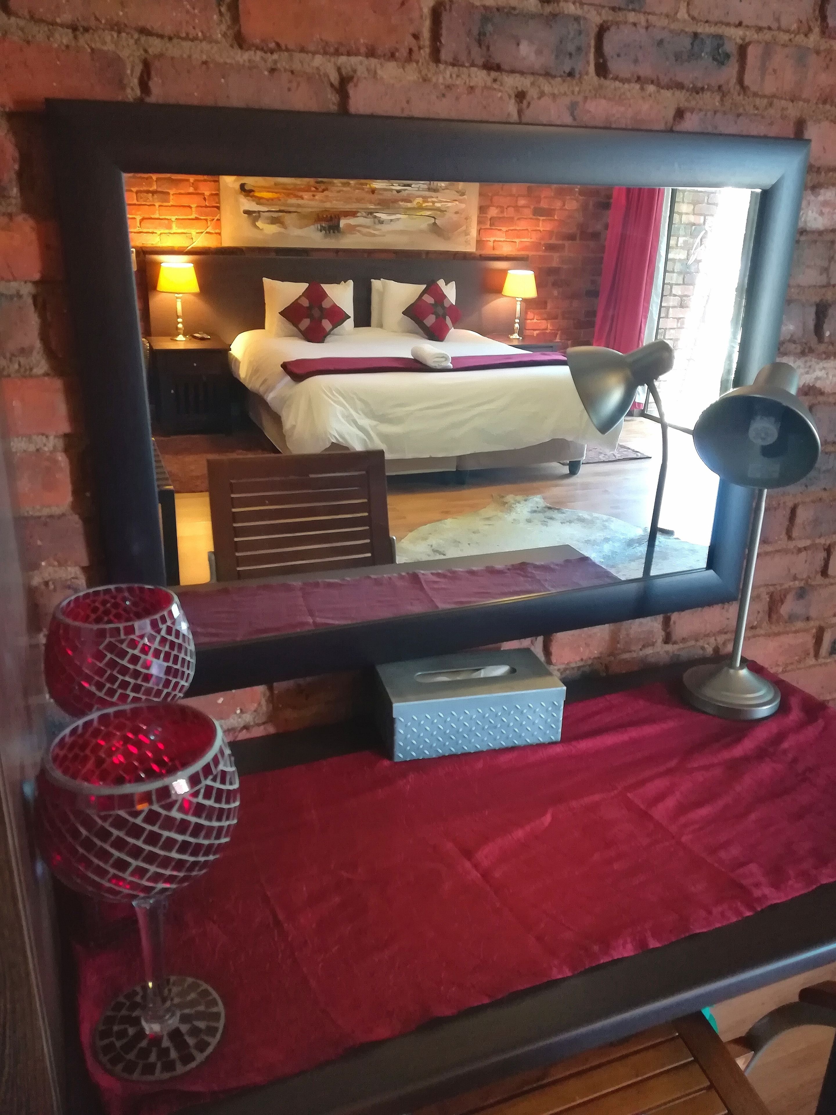 A hotel room with a large mirror reflecting a bed, two bedside lamps, a tissue box, and two decorative red glasses on a red cloth.