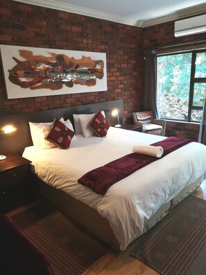 A hotel room with a large bed, a brick wall, a window with a view of trees, and a painting above the bed.