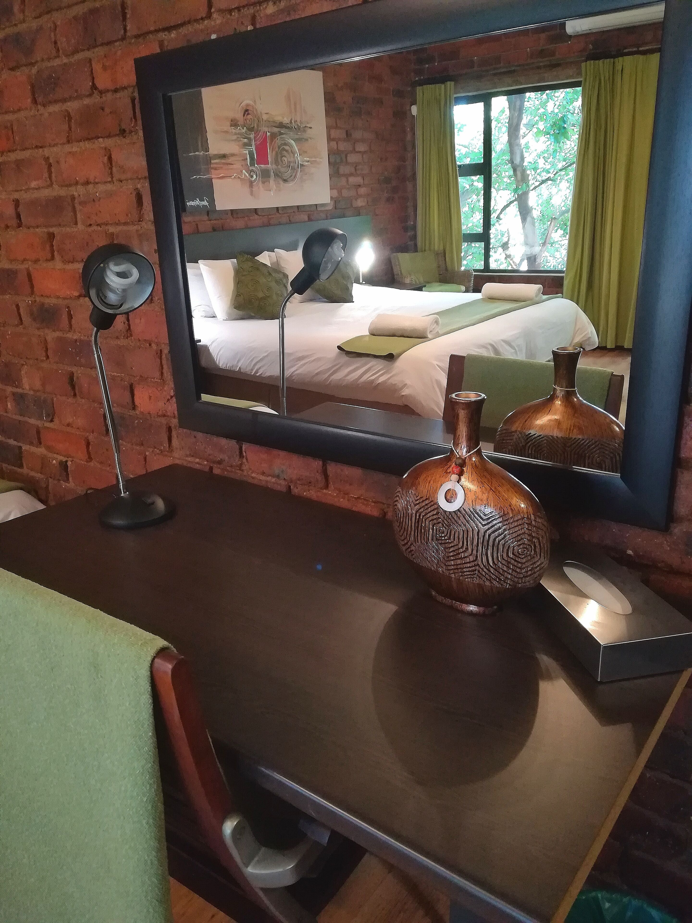 Une chambre d’hôtel avec un mur en briques, un grand miroir qui reflète le lit et la fenêtre, un bureau agrémenté de deux vases décoratifs et une chaise verte.