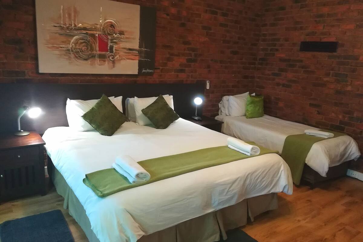 Ein Hotelzimmer mit zwei Betten, jeweils mit weißen Laken und grünen Ziertelementen. Darüber an der Wand hängt ein Gemälde, ein Nachttisch mit einer Lampe und ein Holzboden.
