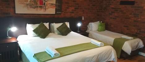 A hotel room with two beds, each with white linens and green throw pillows. There is a painting on the wall above the beds, a nightstand with a lamp, and a wooden floor.