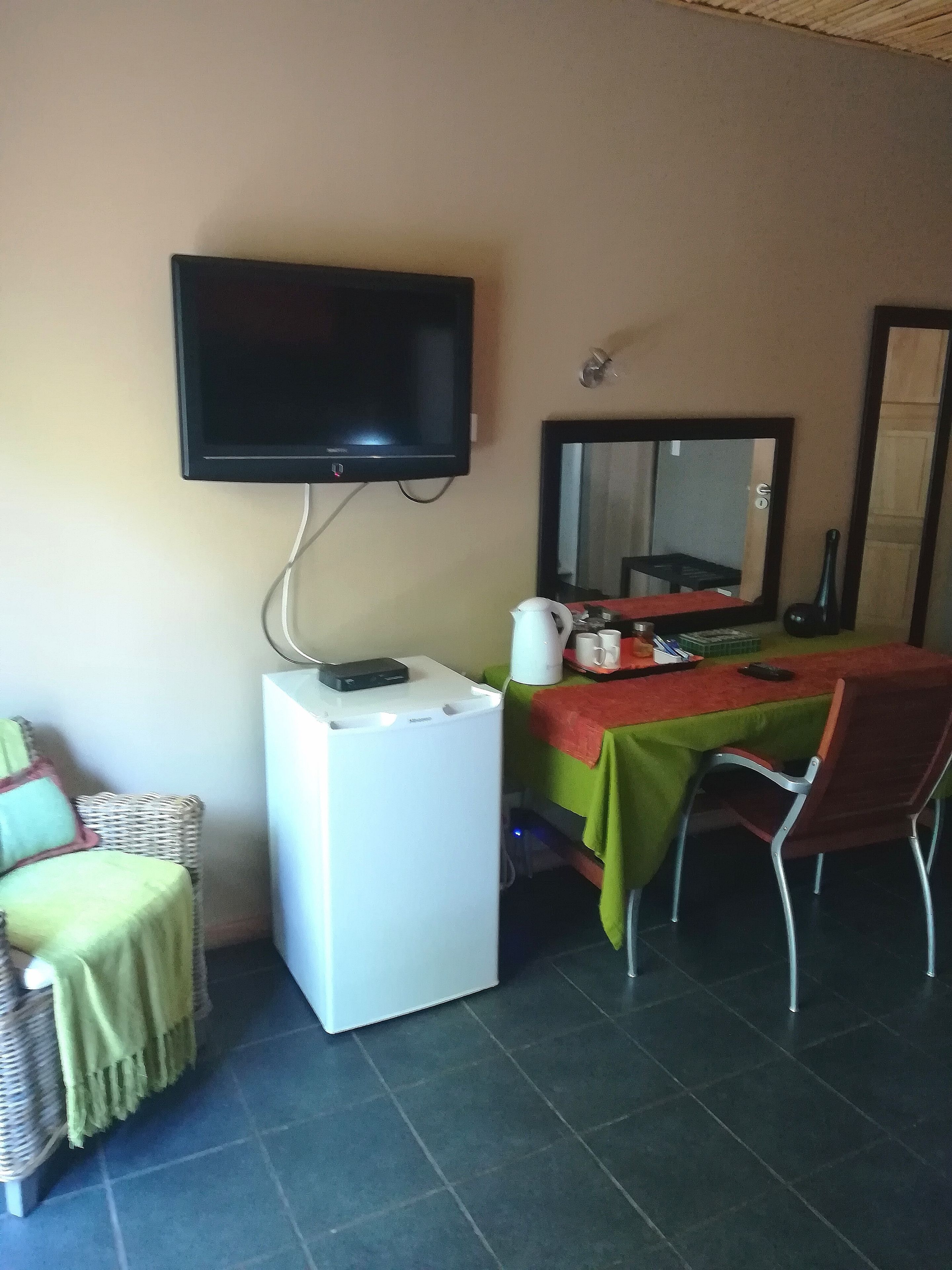 Una habitación con una televisión montada en la pared, un refrigerador blanco pequeño, una mesa con mantel verde y una silla.