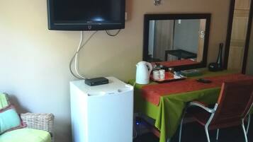 Una habitación con una televisión montada en la pared, un refrigerador blanco pequeño, una mesa con mantel verde y una silla.