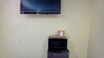 A room with a flat-screen TV mounted on the wall, a black refrigerator, and a microwave on top of it.