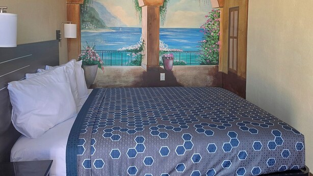 A hotel room with a large bed, a nightstand, a lamp, and a view of the sea through arched windows. - City Center Motel (Indio)