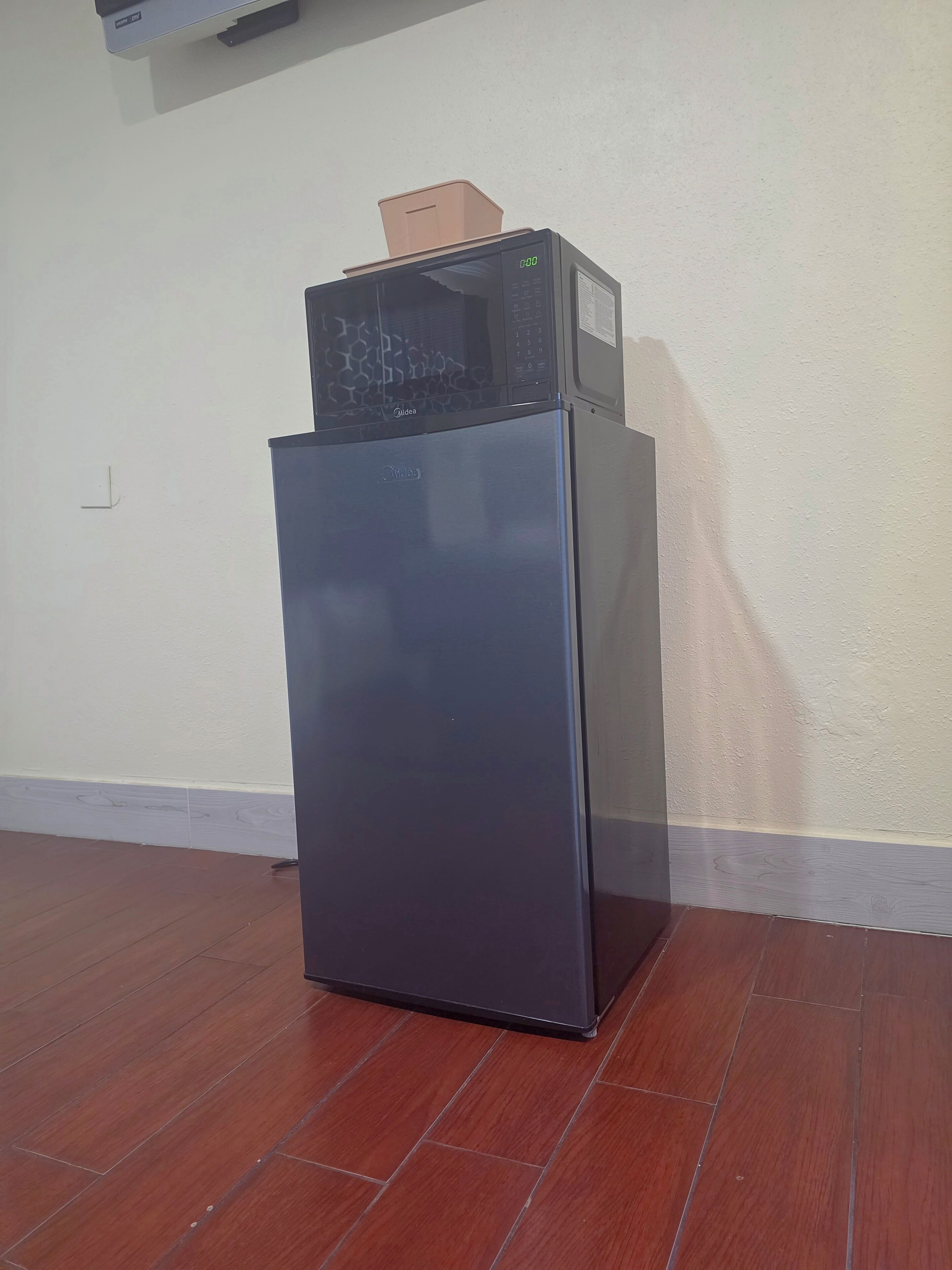 A compact, modern refrigerator with a digital display, placed in a corner of a room with wooden flooring and a plain wall.
