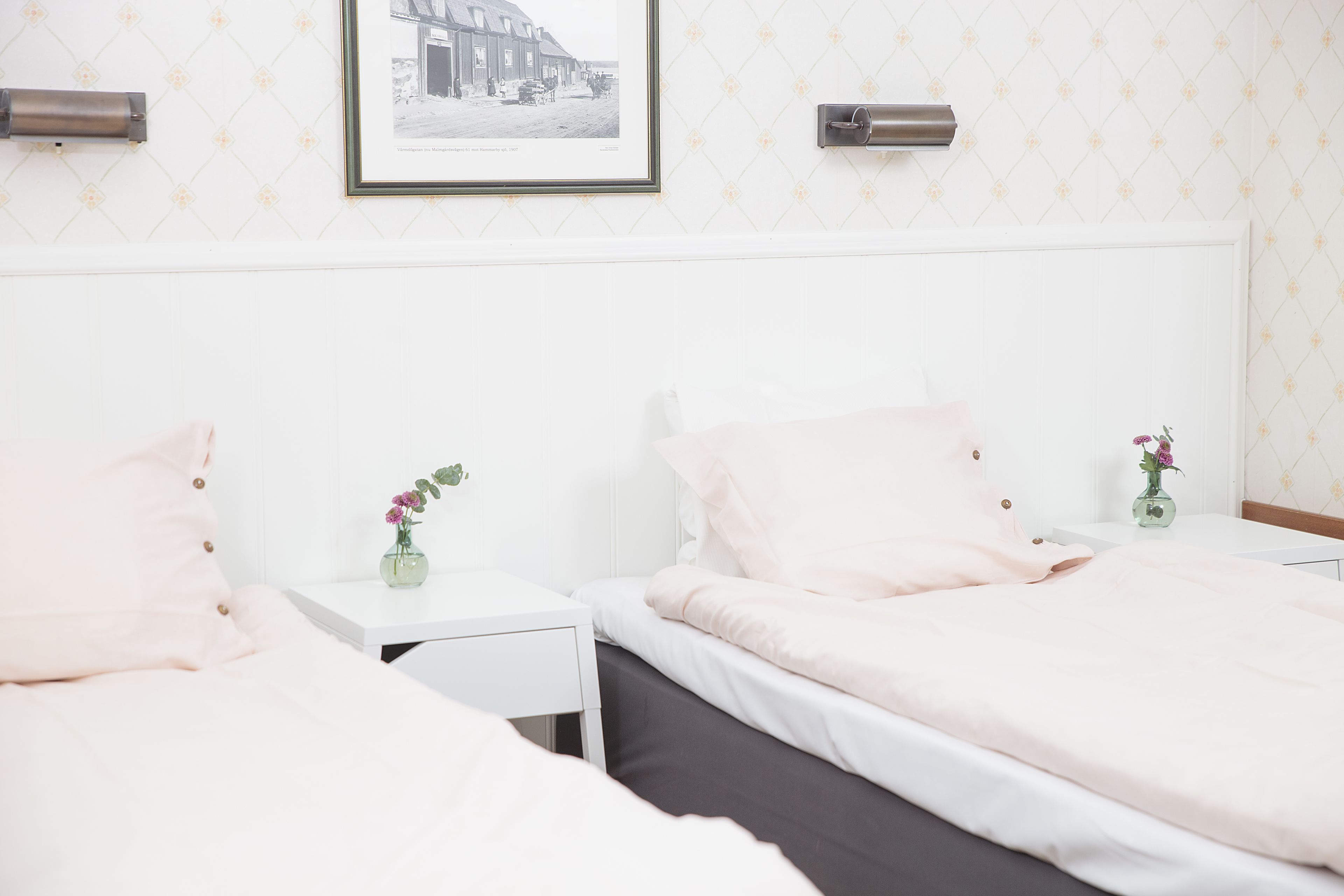 Habitación de hotel con dos camas, paredes blancas, un cuadro enmarcado y un jarrón pequeño con flores.