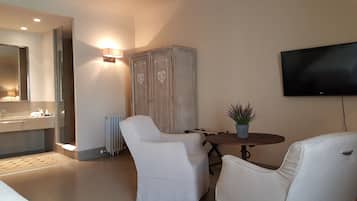 A modern hotel room with a large mirror, a wooden wardrobe, a round table with two white chairs, a potted plant, and a flat-screen TV mounted on the wall.