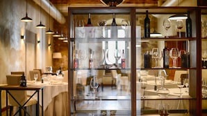 A restaurant interior with wooden beams, hanging pendant lights, and a glass display case with wine glasses and bottles.