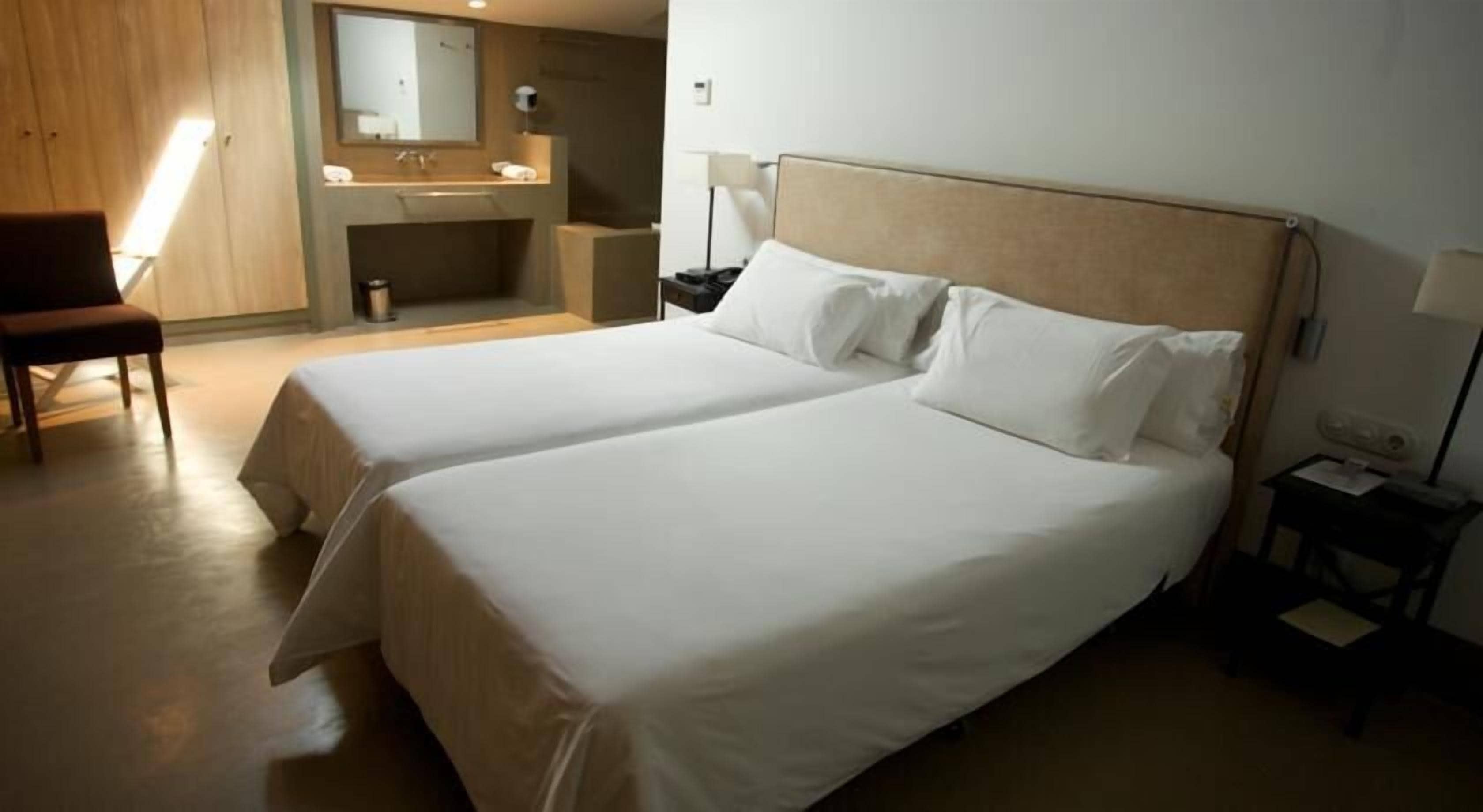 A hotel room with two beds, a desk, a chair, a mirror, and a bathroom with a sink and mirror.