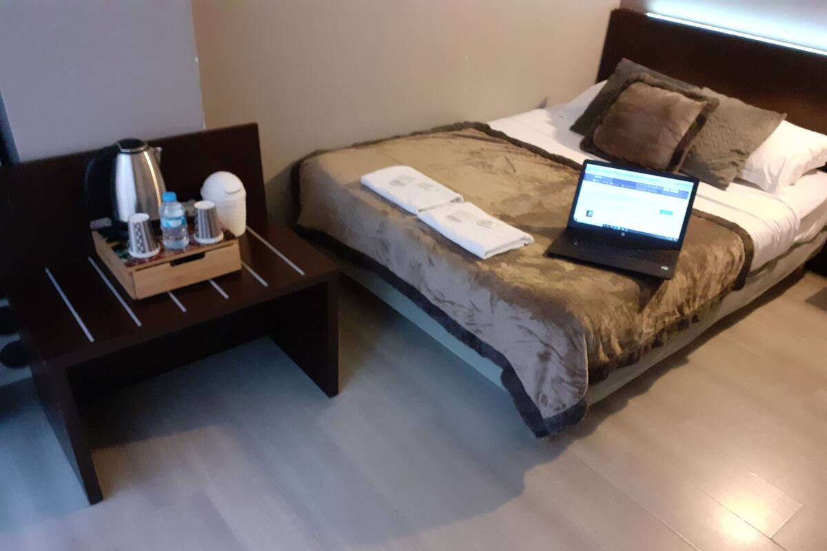 A neatly made bed with a laptop on top, a bedside table with a kettle, cups, and a bottle, and a wooden bench beside the bed.