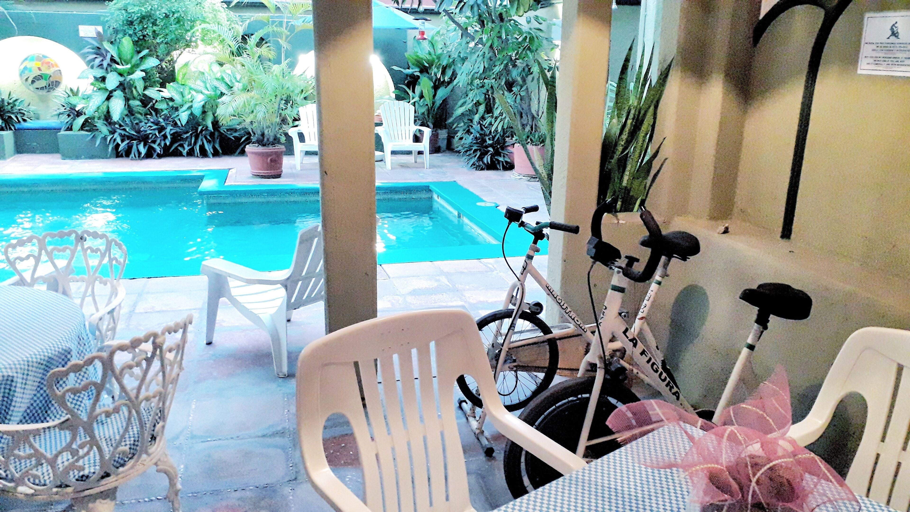 Un área junto a la piscina con sillas de plástico blancas, una bicicleta y plantas en macetas.