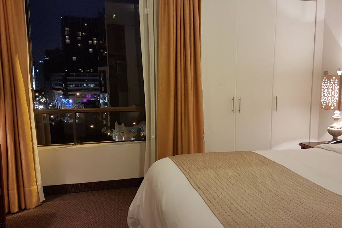 A hotel room with a bed, a nightstand with a lamp, a window with curtains, and a view of the city at night.