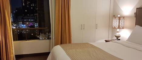 A hotel room with a bed, a nightstand with a lamp, a window with curtains, and a view of the city at night.
