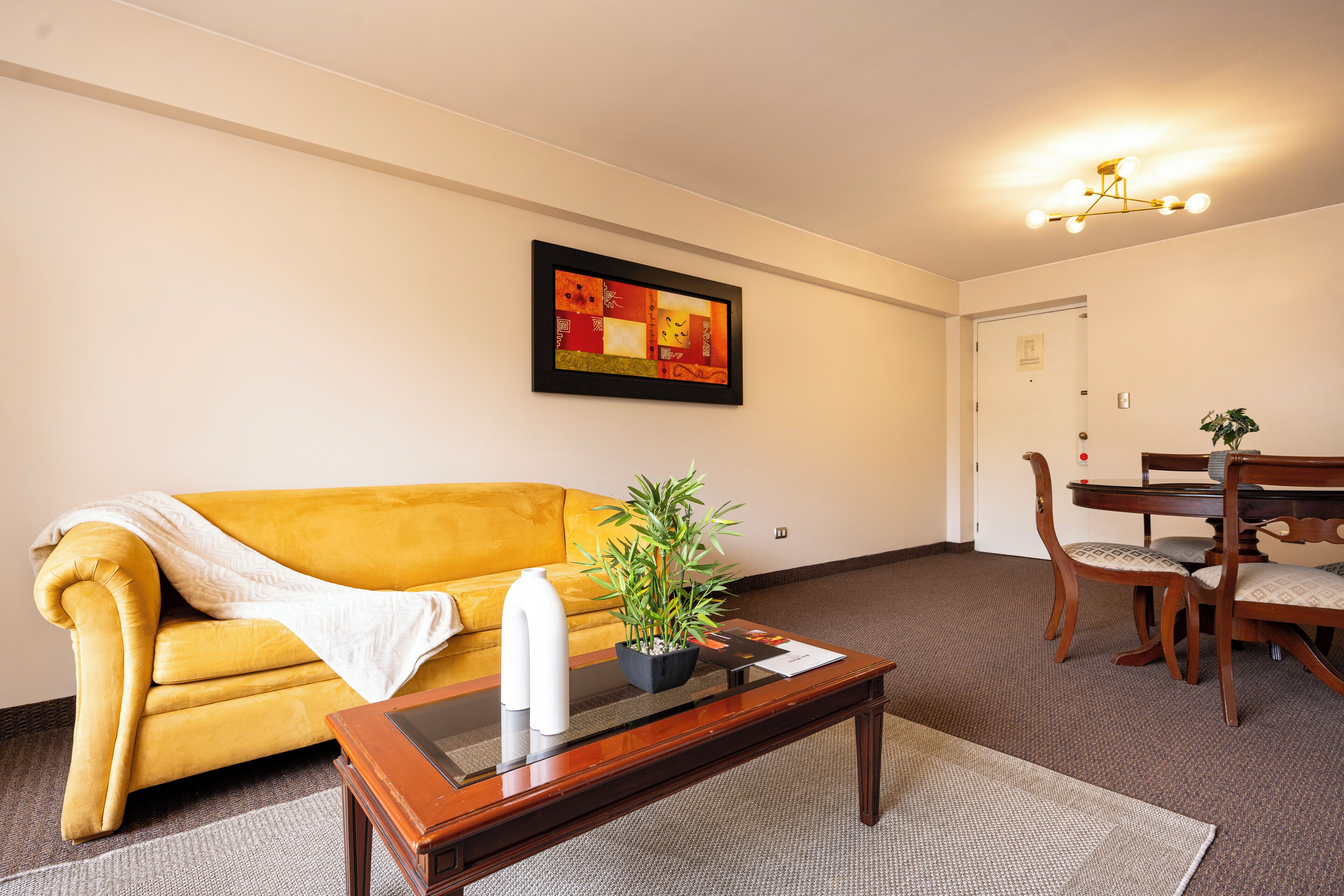 A living room with a yellow sofa, a coffee table with a plant, a framed artwork, and a dining area with a wooden table and chairs.