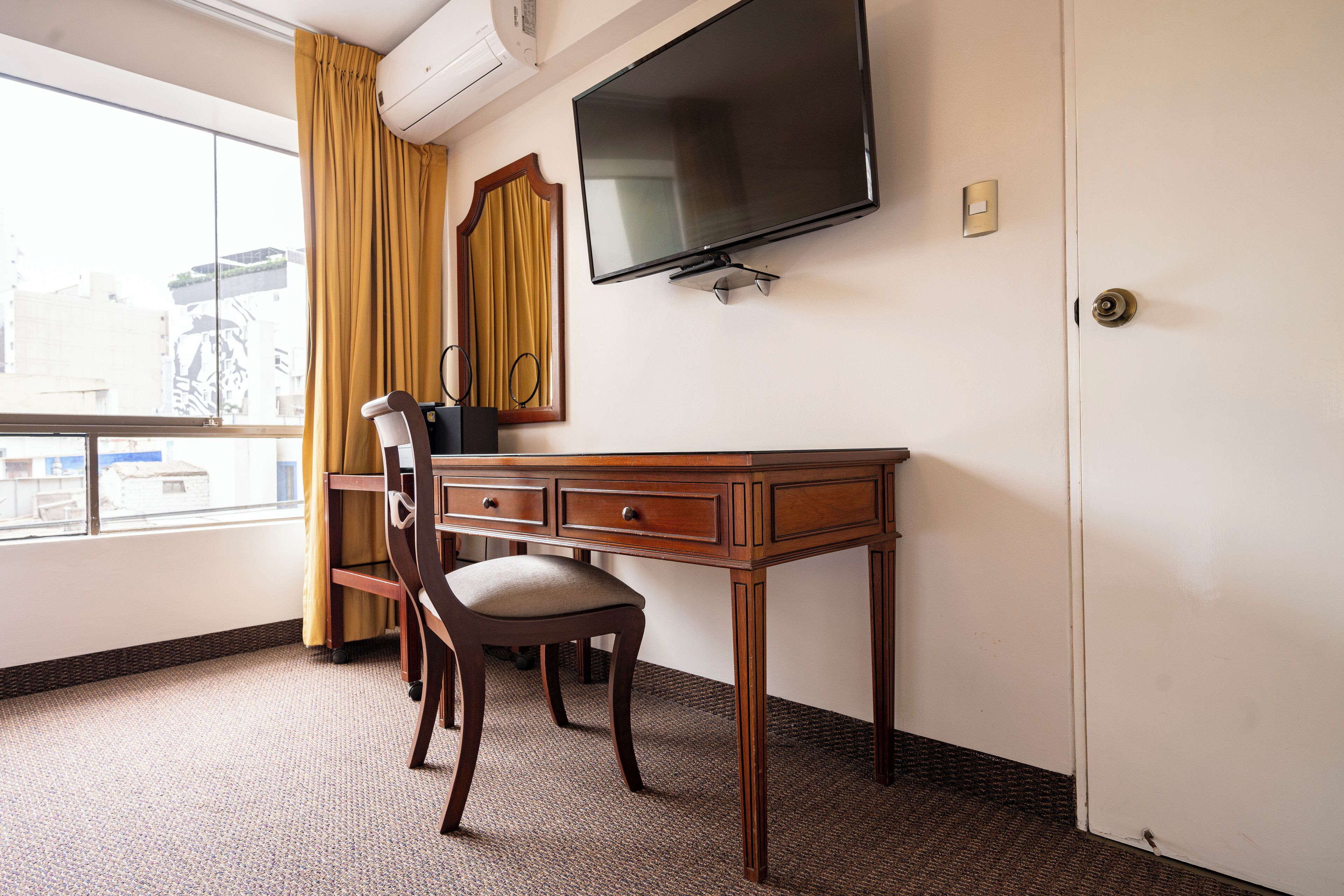 A hotel room with a wooden desk, a chair, a flat-screen TV mounted on the wall, and a window with yellow curtains.