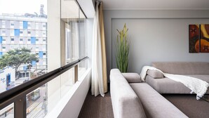 A modern living room with a sofa, a tall plant, and a view of a cityscape through a large window. - Apart Hotel San Martin (Lima)