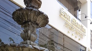 A stone fountain with water cascading down, surrounded by a circular stone basin, in front of a building with a sign reading "San Antonio Hotel". - Apart Hotel San Martin (Lima)