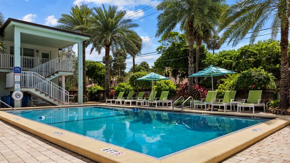 A swimming pool with lounge chairs and umbrellas, surrounded by palm trees and a building with a balcony.