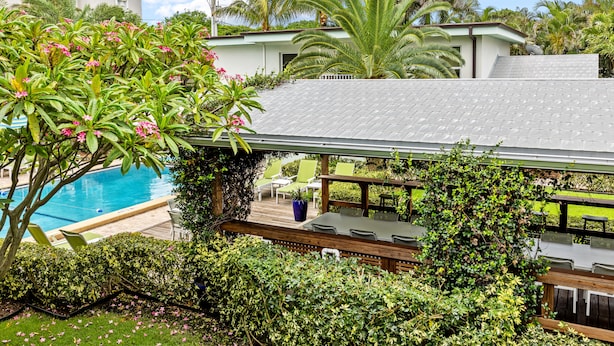 A poolside area with a covered seating area, lush greenery, and a view of a building with a tiled roof. - Sun Dek Beach House (Boynton Beach)