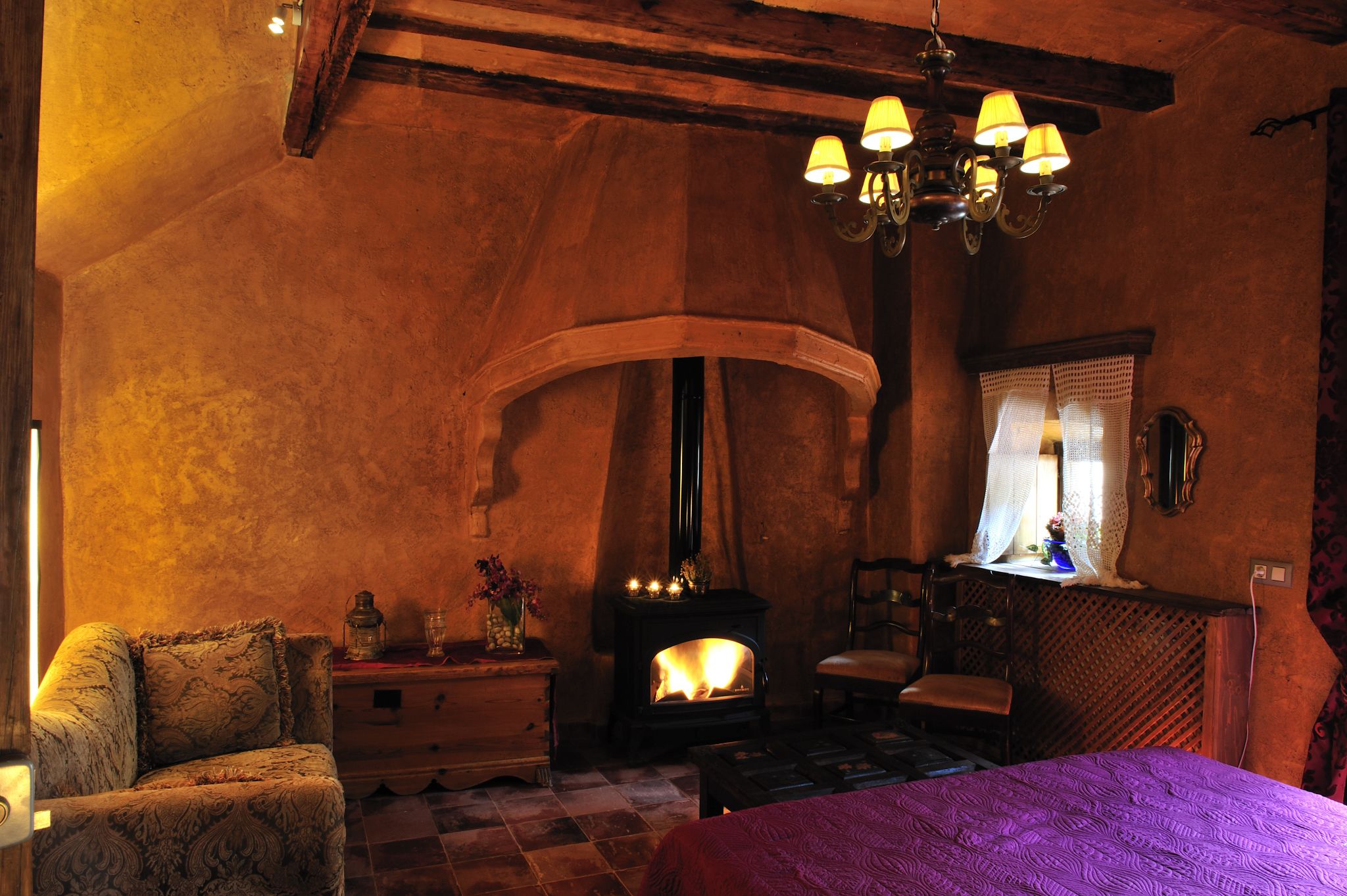 Una stanza accogliente con camino, una sedia in legno, un letto con copriletto viola e un lampadario.