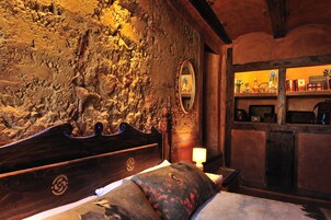 Una habitación rústica con pared de piedra, cama de madera y estantes con libros y objetos decorativos.