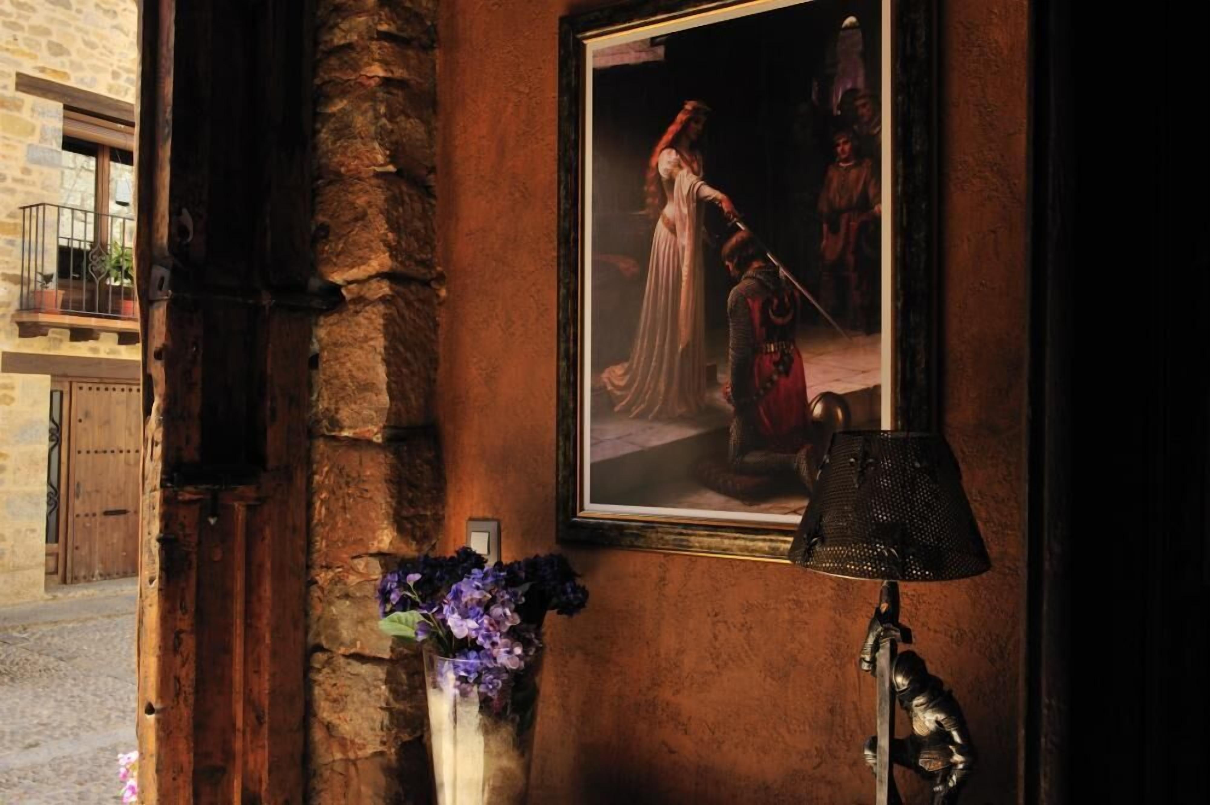 A rustic interior with a stone wall, a framed painting, a vase with flowers, and a lamp.