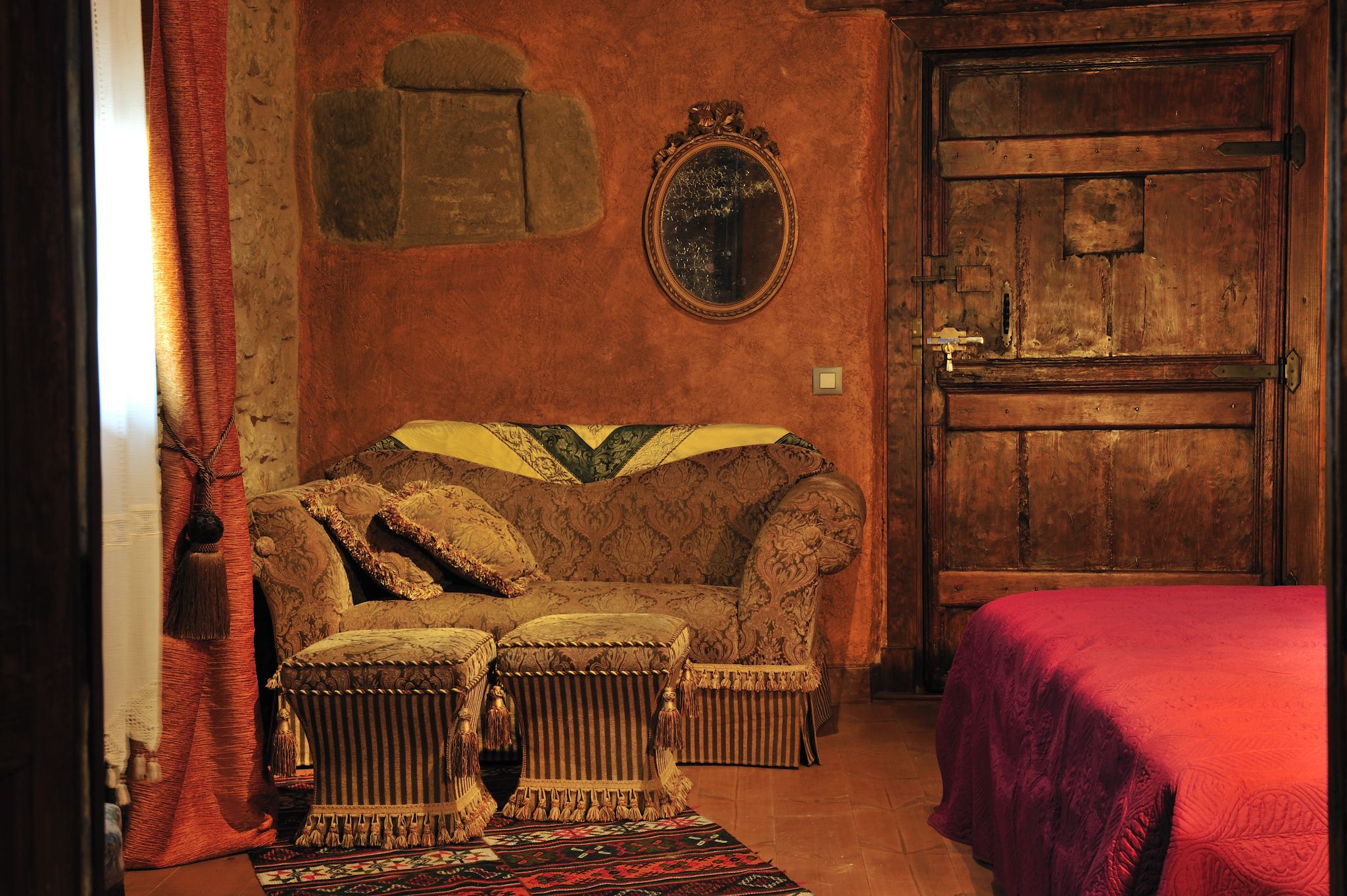 A room with a vintage sofa, two ottomans, a red bedspread, a wooden door, and a mirror on the wall.
