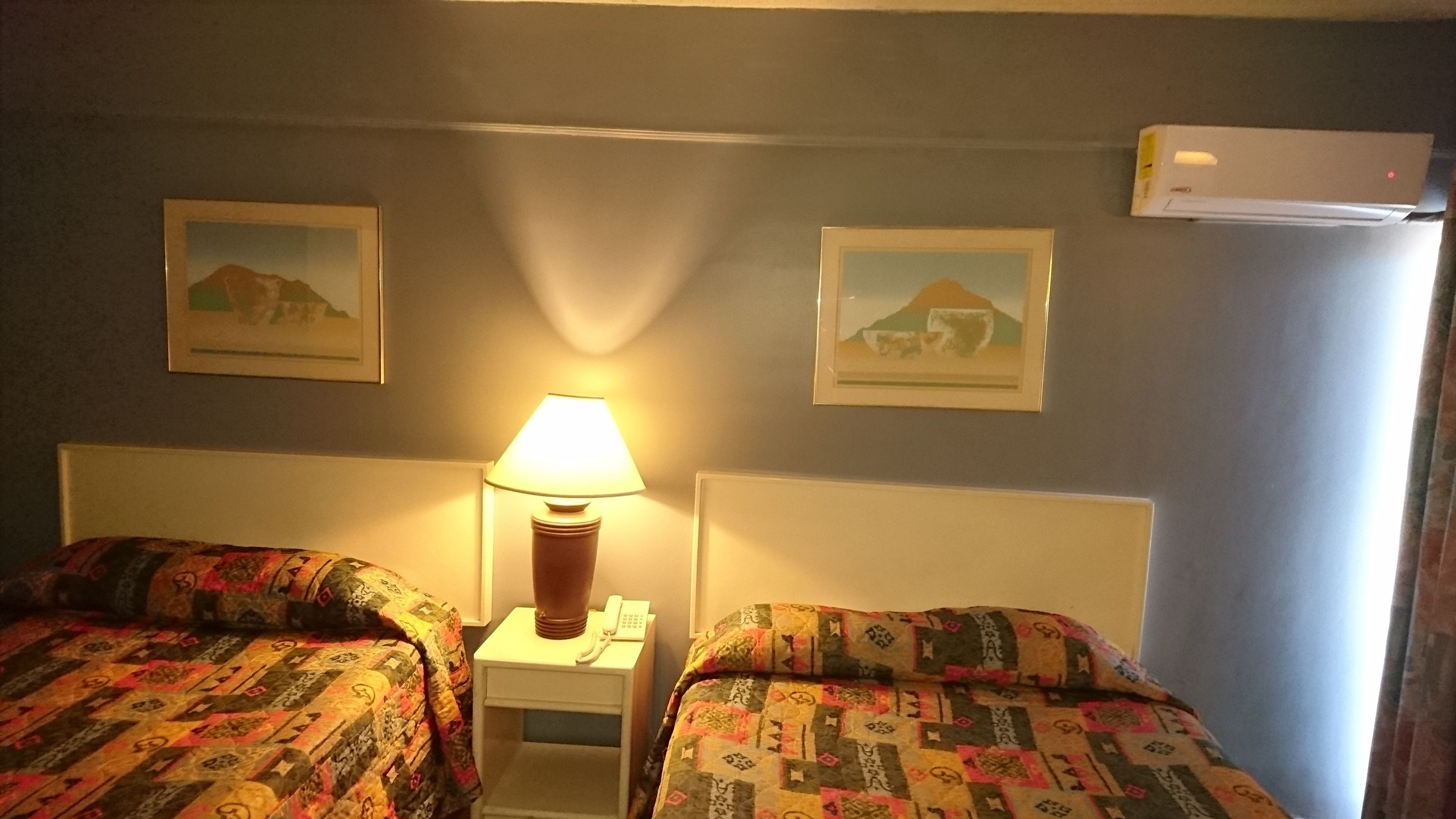 Habitación de hotel con dos camas, una mesita de noche con lámpara, y dos cuadros enmarcados en la pared.