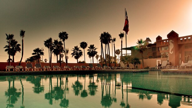 A swimming pool with lounge chairs and palm trees, reflecting in the water.