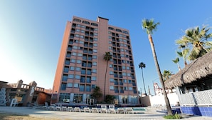 A tall hotel with multiple balconies, a swimming pool area with lounge chairs, and palm trees.