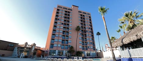 Un hotel alto con varios balcones, una zona de piscina con sillones de descanso y palmeras.