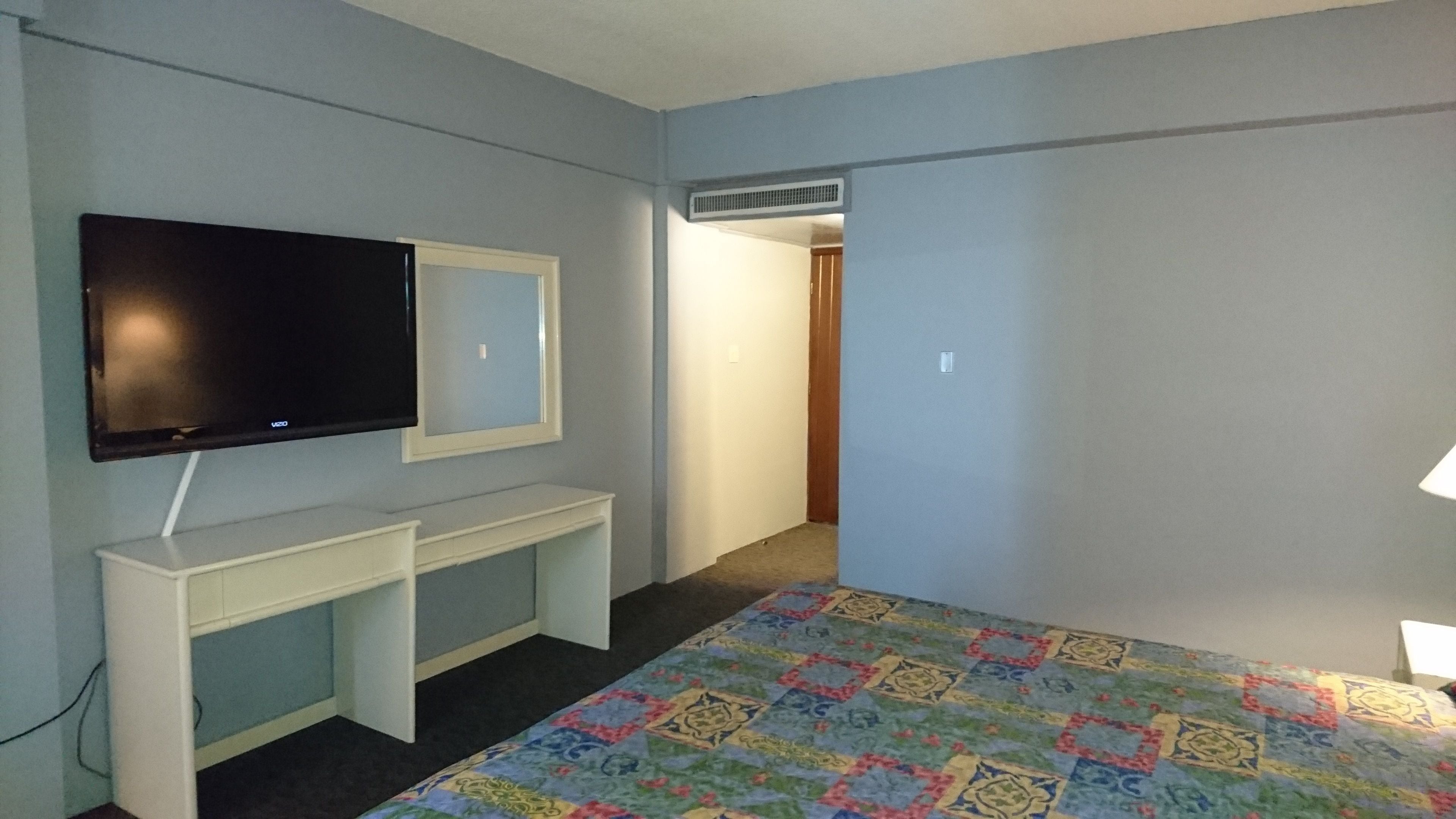 A bedroom with a bed, a TV mounted on a wall, a desk, and an air conditioning vent.