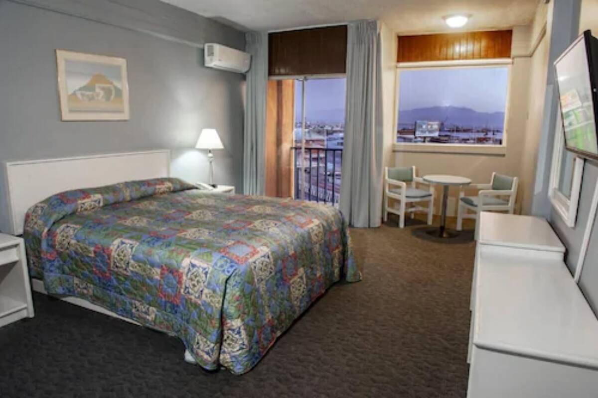 Une chambre d’hôtel comprenant un lit, un bureau, une chaise, une petite table et une fenêtre donnant sur un paysage urbain et des montagnes.