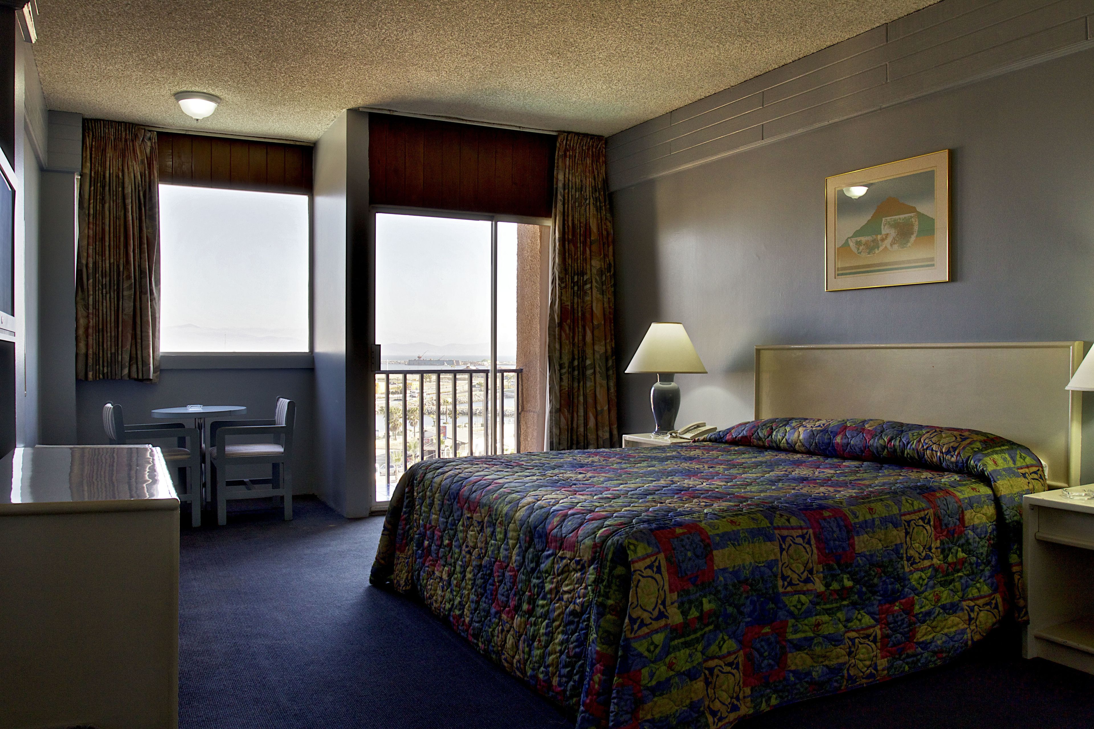 A hotel room with a large bed, a desk, and a chair. There is a window with curtains, a lamp on the bedside table, and a painting on the wall.
