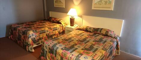 A hotel room with two beds, each covered with a patterned quilt. There is a bedside lamp on one of the beds, and two framed pictures on the wall.