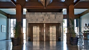 A spacious lobby with a polished marble floor, a large stone wall, and a wooden ceiling. There are two large potted plants and a reception desk with chairs.