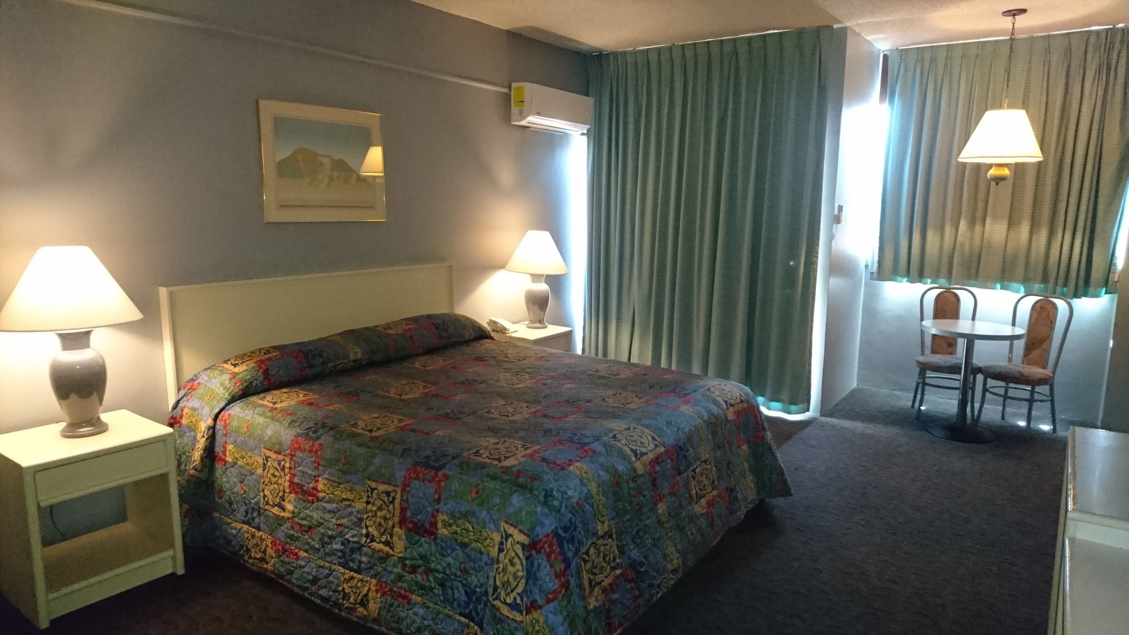 A hotel room with a bed, two bedside lamps, a desk with a chair, and a window with curtains.