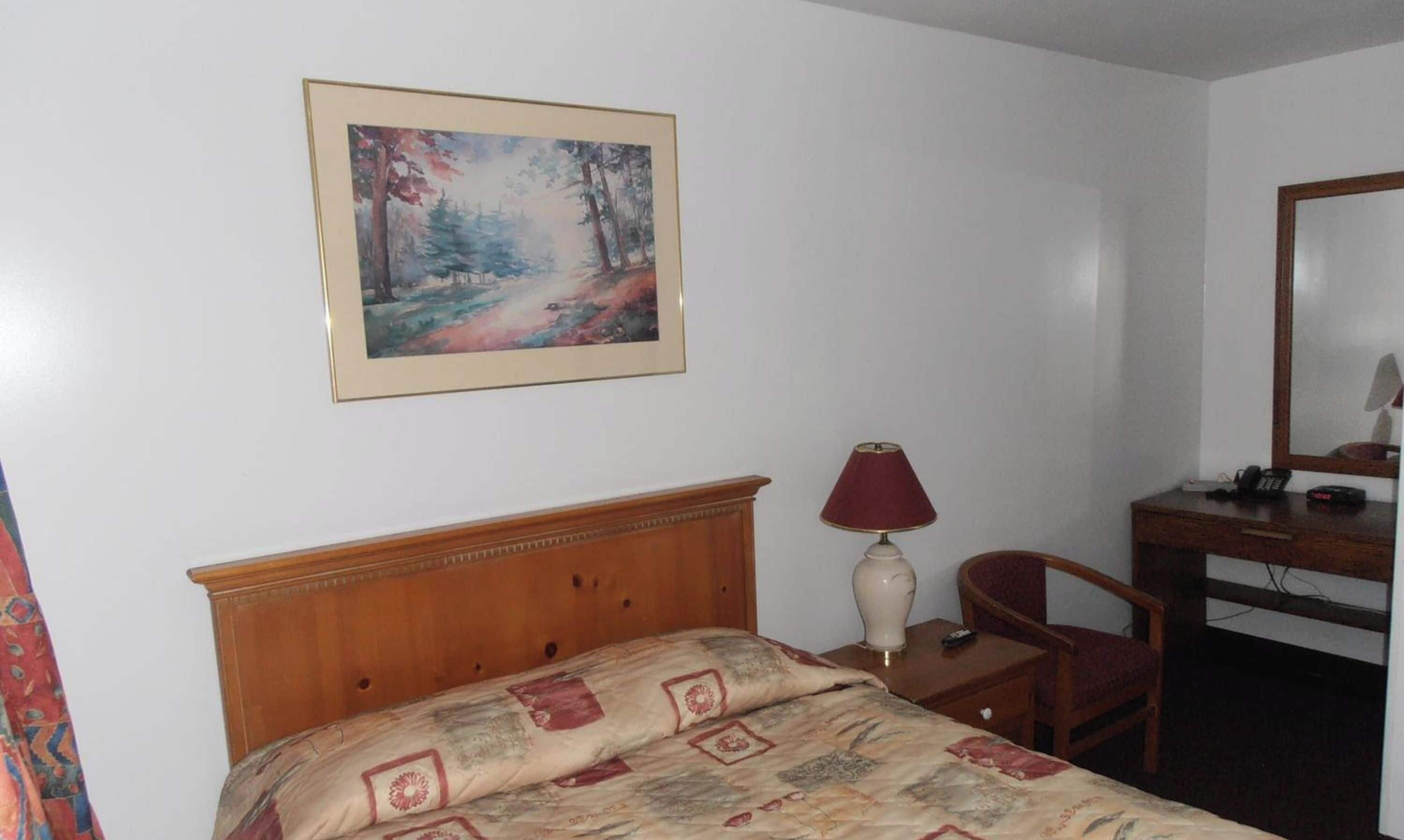 Habitación de hotel con cama, escritorio con lámpara, una silla y un cuadro en la pared.