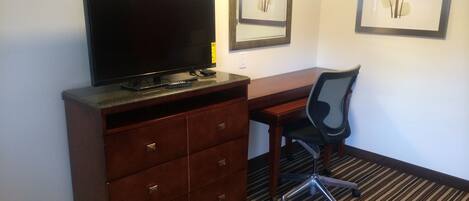 A hotel room with a desk, chair, television, and framed artwork on the wall.