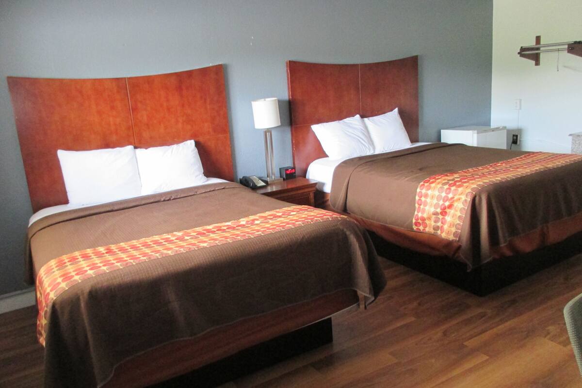 Two double beds with brown headboards and white pillows, a bedside table with a lamp, and a wooden floor.