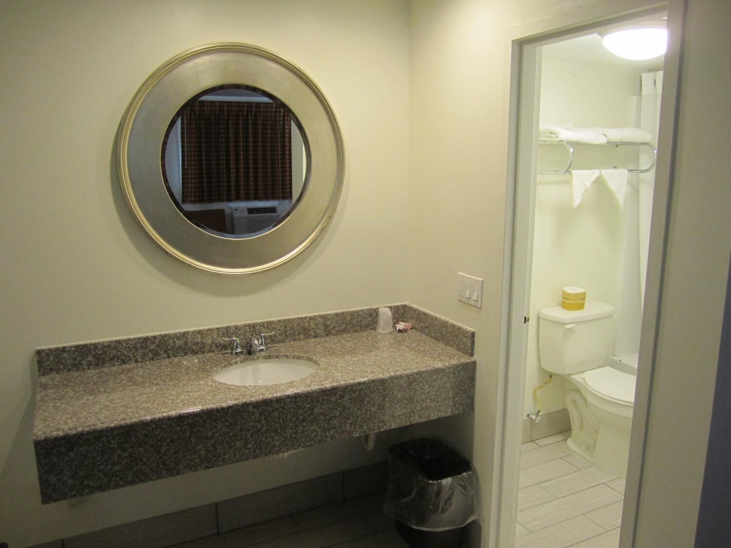 A bathroom with a granite countertop sink, a round mirror, and a toilet visible through an open door.