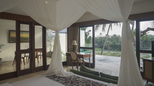 A bedroom with a large bed, a desk, and a view of a pool and tropical landscape.