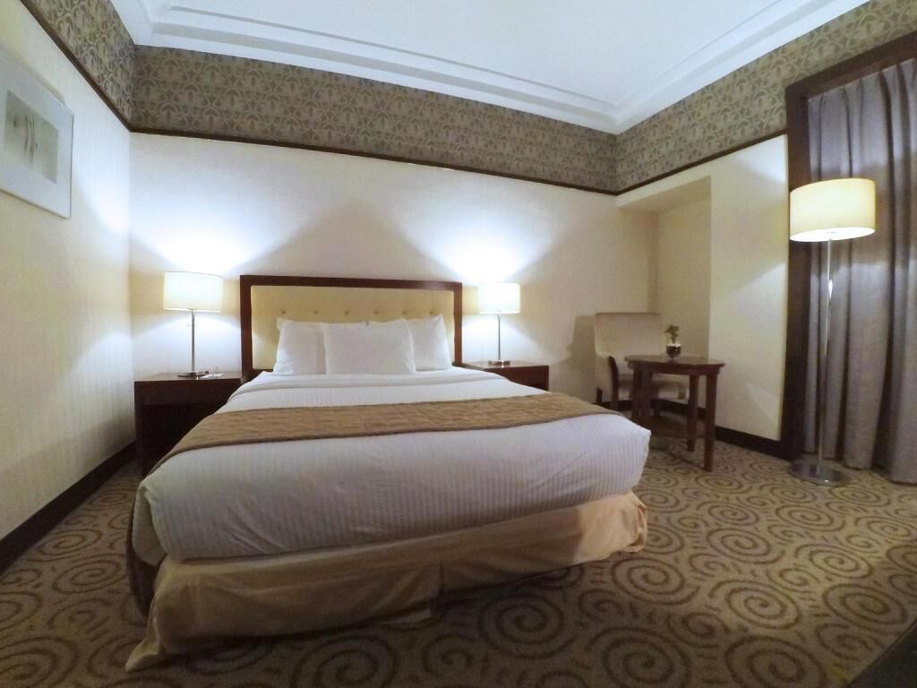 A hotel room with a large bed, two bedside tables with lamps, a small table, and a curtain.