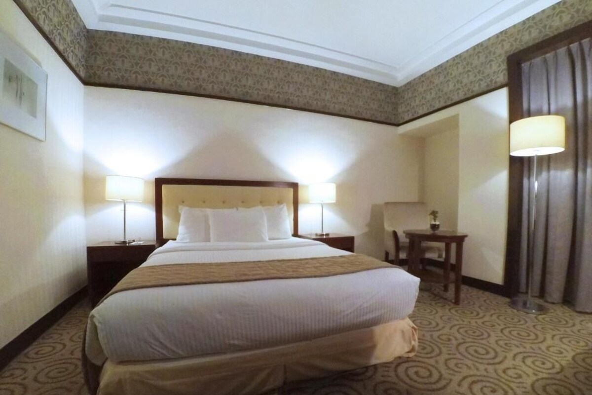 A hotel room with a large bed, two bedside tables with lamps, a small table, and a curtain.
