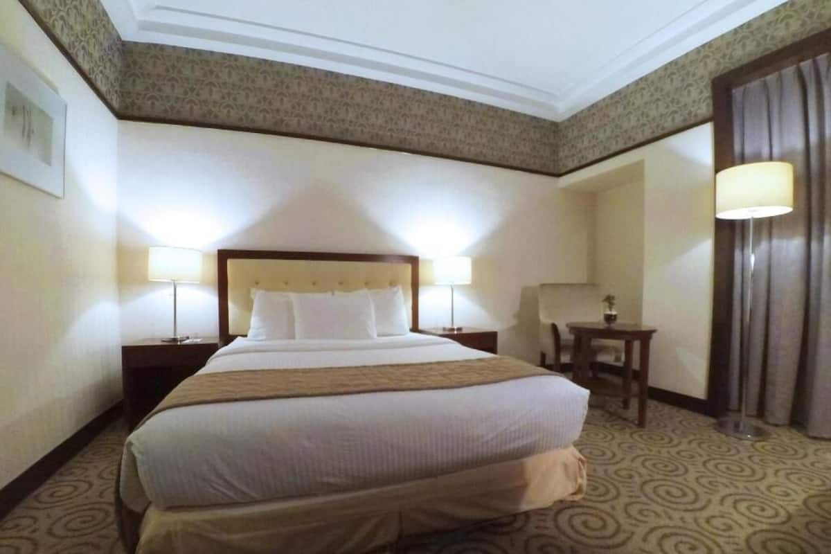 A hotel room with a large bed, two bedside tables with lamps, a small table, and a curtain.