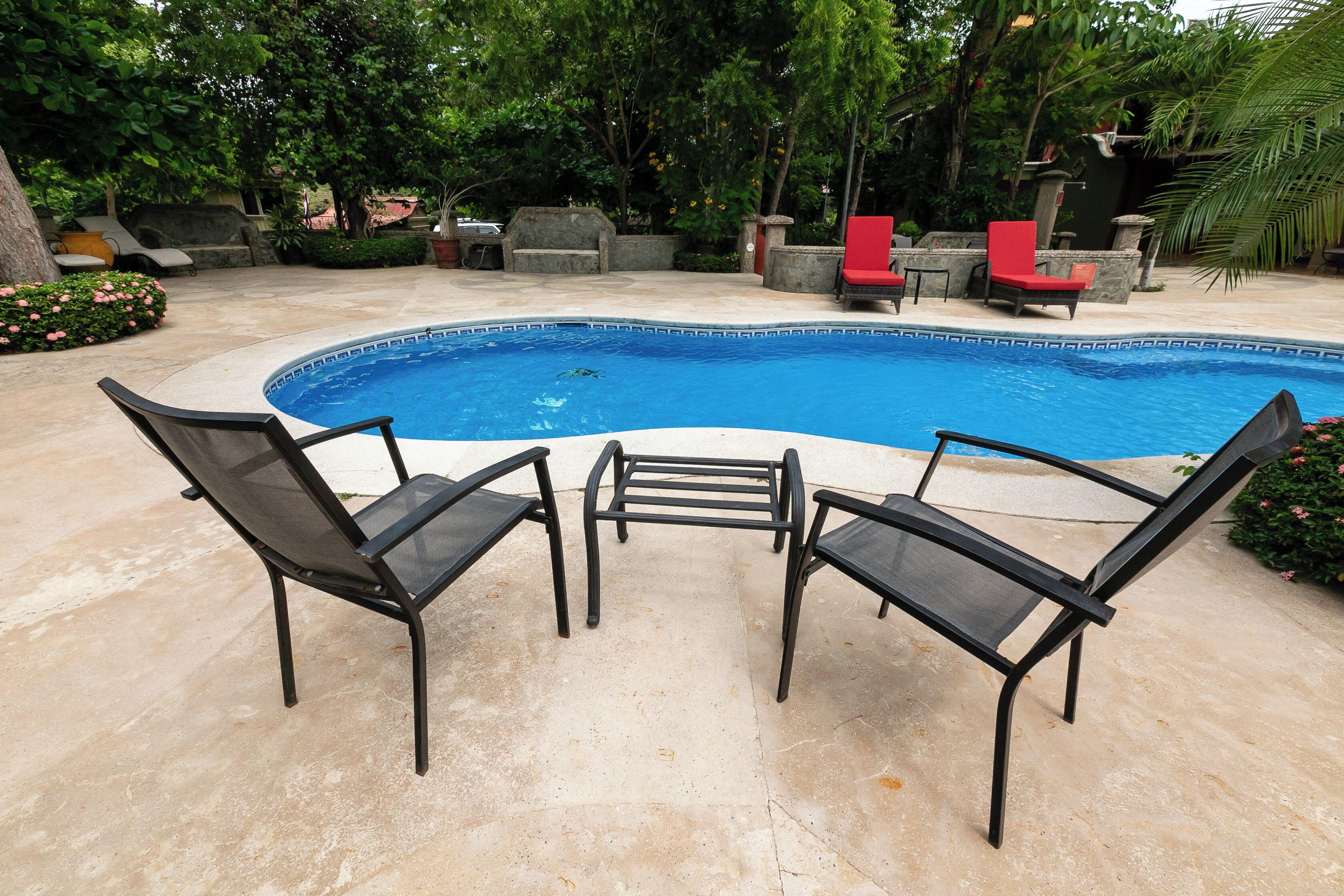 Ein Swimmingpool mit zwei schwarzen Liegestühlen und einem roten Sessel, umgeben von Grünpflanzen und Steinbauten.