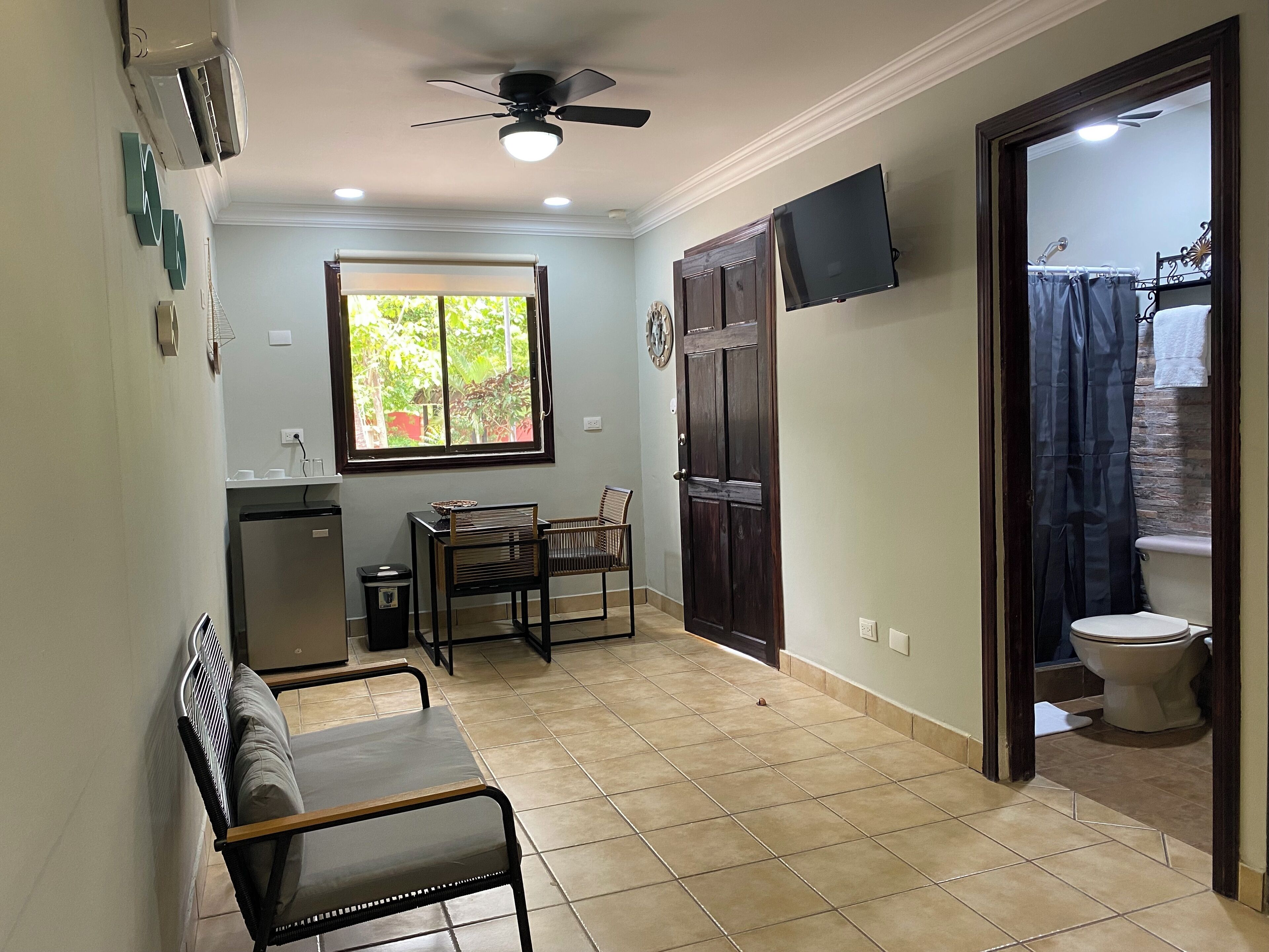 Habitación compacta con mesa de comedor, sillas, un refrigerador pequeño, ventilador de techo, ventana, puerta que da a un baño y televisor montado.