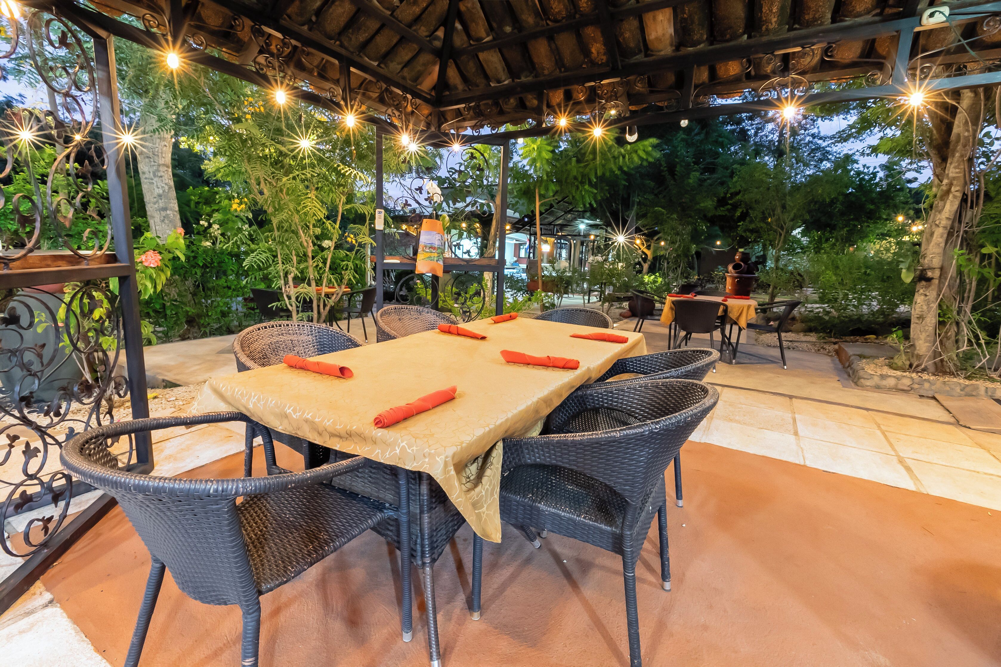 Un área de comedor al aire libre con una pérgola cubierta, sillas de mimbre y una mesa preparada para cuatro comensales.